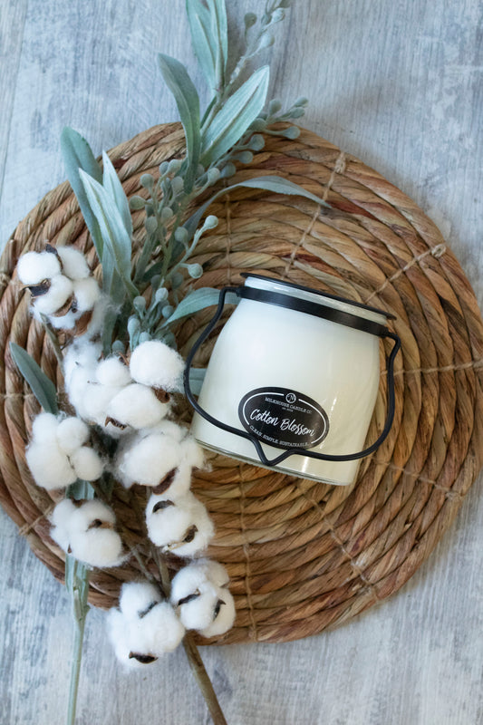 A jar of Milkhouse Candle Company's Creamery Collection Butter Jar with a cotton blossom fragrance, placed on a woven placemat with cotton blossoms and greenery. 